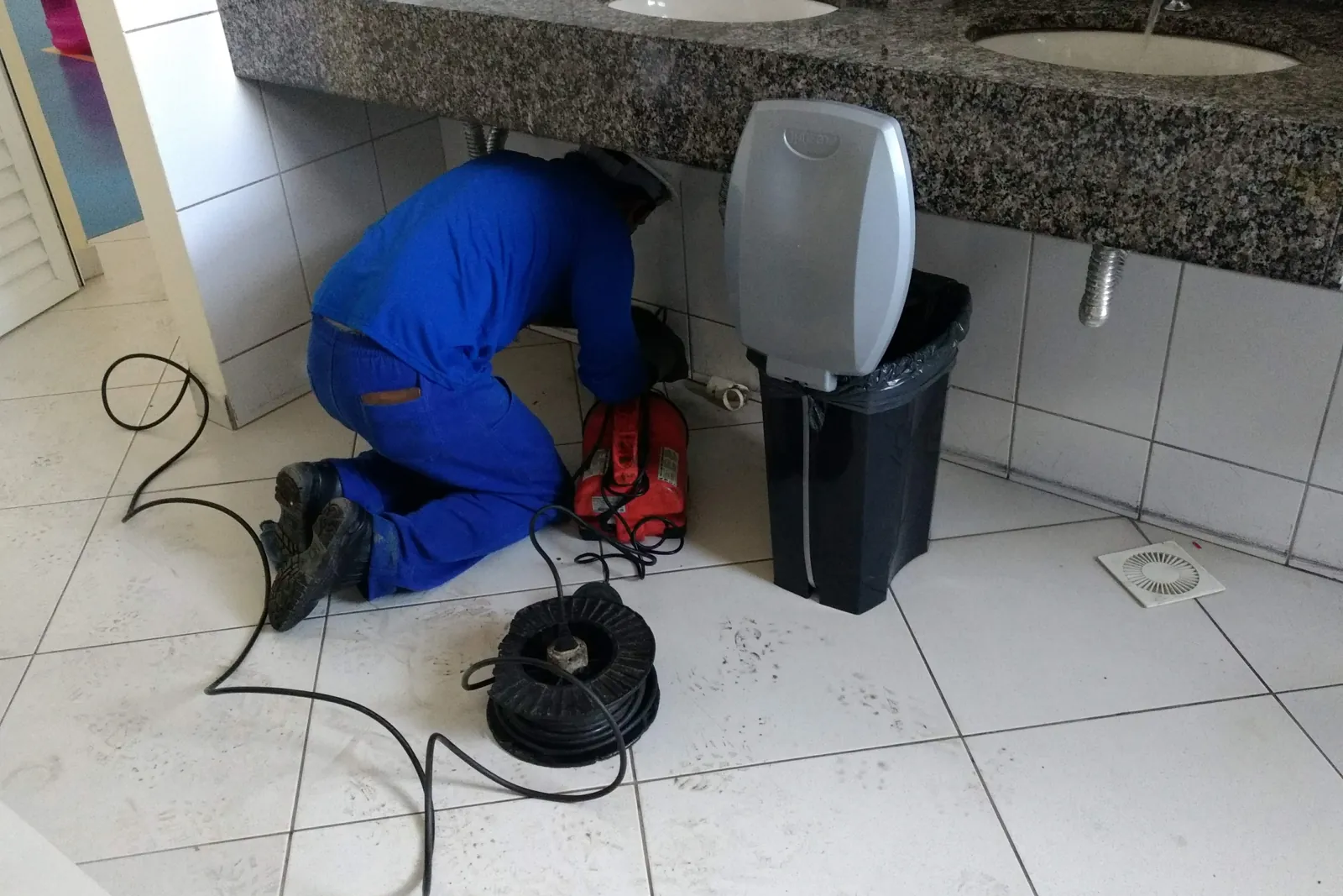 Técnico desentupindo a pia de um banheiro com máquina de cabo, ajoelhado embaixo da bancada